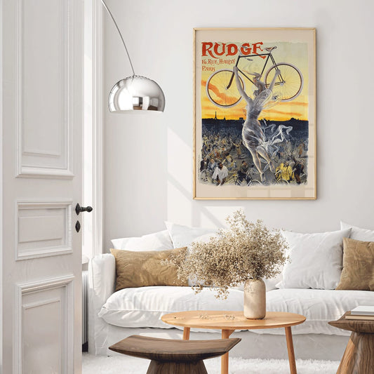 Vintage bicycle poster by Jean de Paleologue showing a triumphant figure and a woman holding a bicycle above a crowd at sunset for Rudge bicycle brand, framed and displayed in home decor